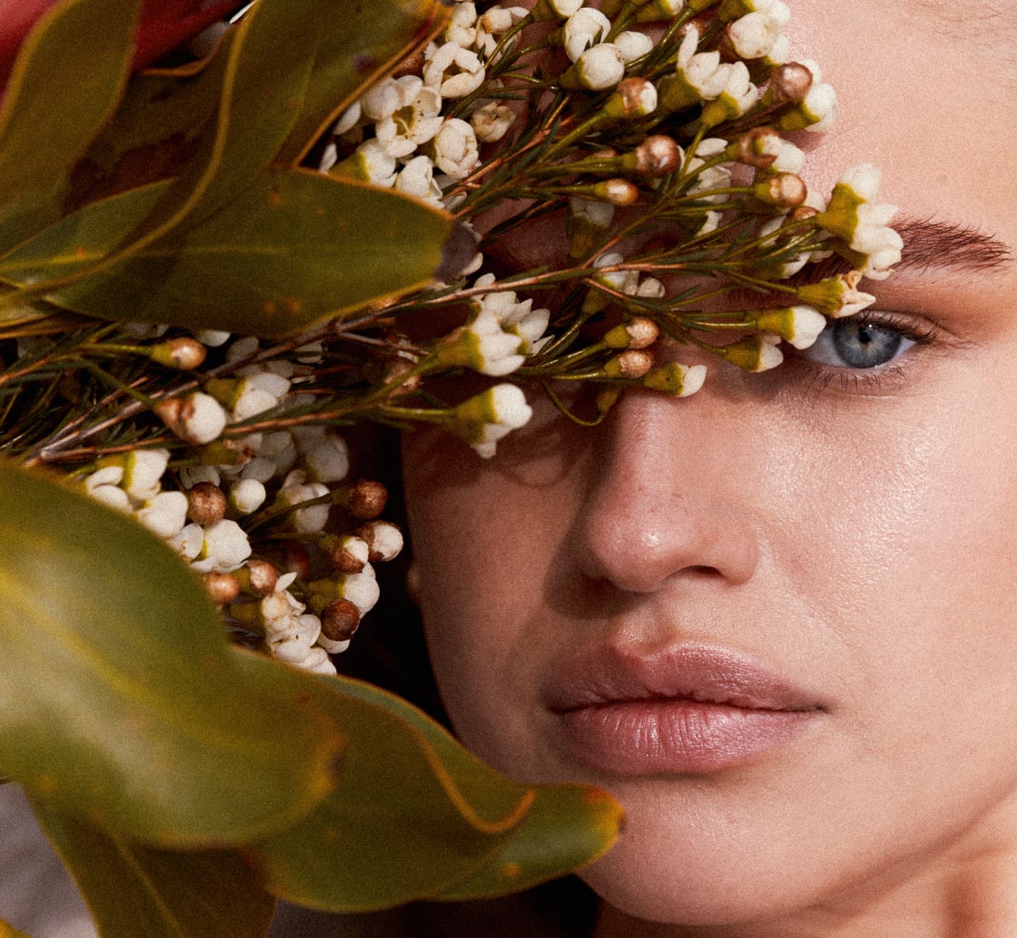 Close-up beauty portrait with natural skin, framed by white blossoms and green foliage, evoking a fresh, botanical aesthetic.