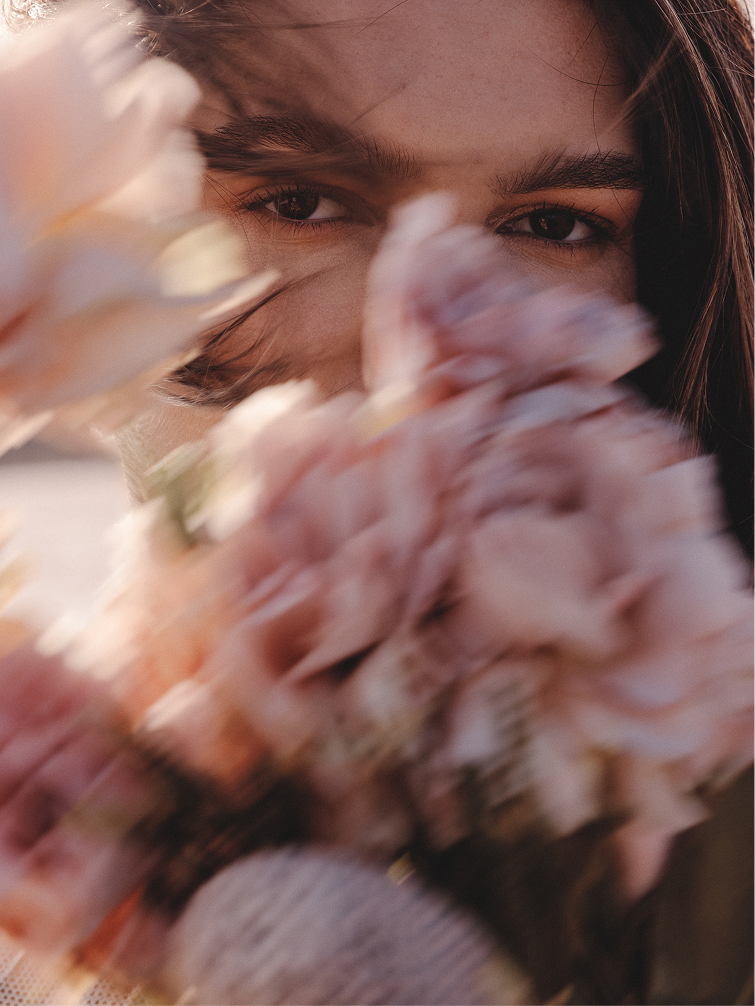 Close-up portrait of a person with soft natural light, partially obscured by a bouquet of pink flowers in the foreground, creating a dreamy, blurred floral effect with focus on the eyes